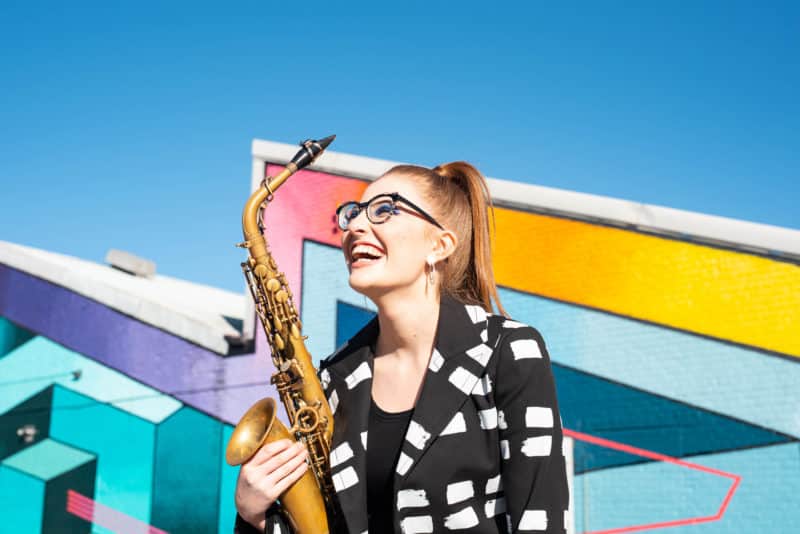 Picture shows Jess Gillam holding a saxophone against a colourful building and a bright blue sky - London Classical Music Concert